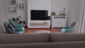 Een woonkamer met een grijze bank, twee turquoise fauteuils, een flatscreen televisie op een witte standaard, en een kast met decoratieve objecten.