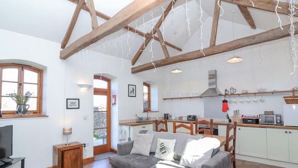 A cozy living room with a grey sofa, wooden beams, a kitchen with white cabinets, and a TV. - The Yarde Annexe (Taunton)