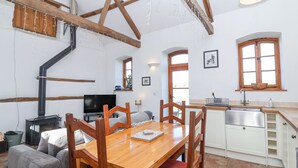 A cozy living room with a wooden dining table, red cushioned chairs, a sofa, and a TV. There is a kitchen area with a sink, cabinets, and a window. - The Yarde Annexe (Taunton)