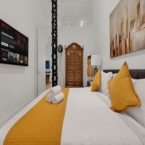 A hotel room with a bed, a wooden wardrobe, a wall-mounted TV displaying a Netflix show, and a painting on the wall.