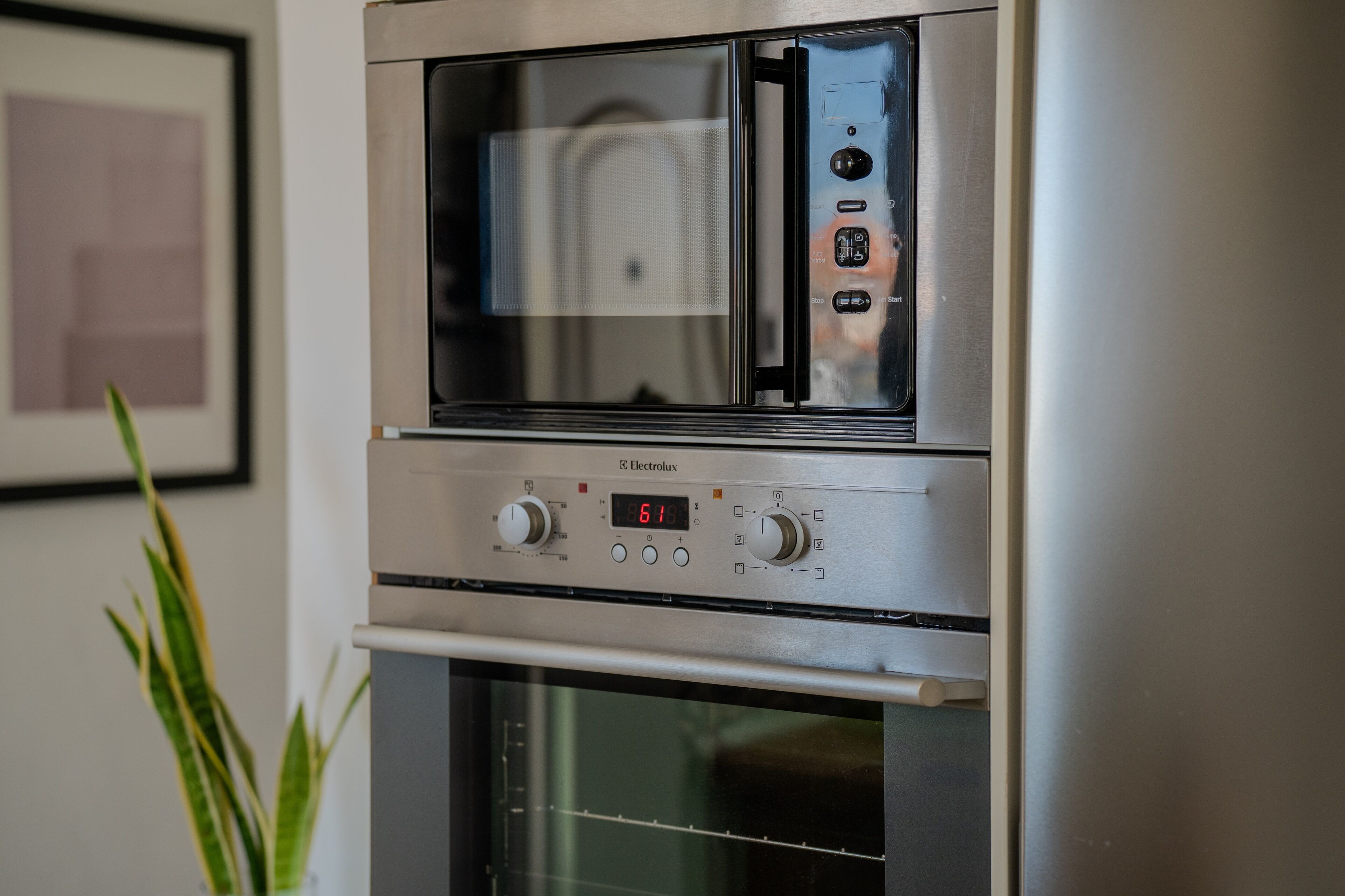 A stainless steel oven with a digital display and control knobs, situated in a kitchen with a framed picture on the wall.