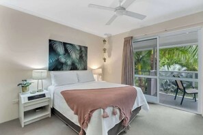 Una camera da letto con un letto, una ventola a soffitto, un comodino con una lampada, un'ampia finestra vista palme e un balcone con sedie.