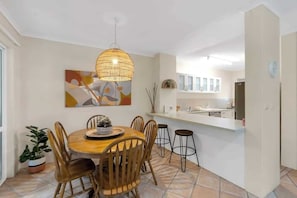 A dining area with a round table, wooden chairs, a hanging light fixture, and a kitchen in the background.