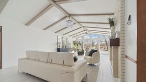 A spacious living area with a high ceiling, a white sofa, a coffee table, and a view of the outdoors through large windows. - Waterfront Paradise - 2 Lake Views in Prime Location (Battle Creek)