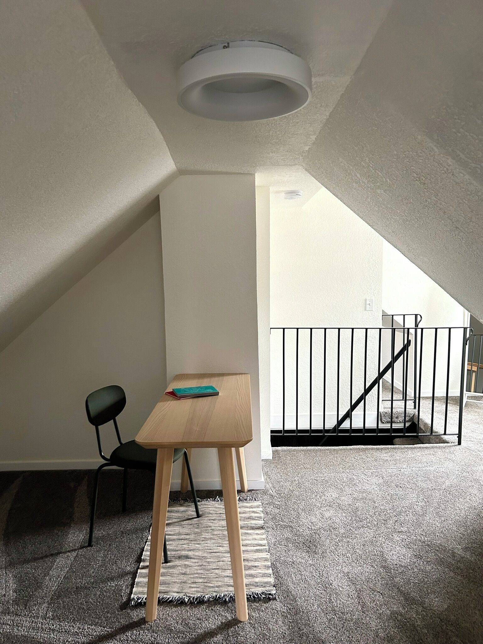 Una habitación pequeña y bien iluminada, con techo inclinado, una mesa de madera, una silla negra y una barandilla de metal negro.