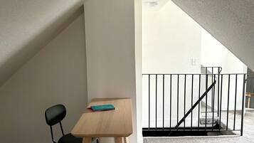 Una habitación pequeña y bien iluminada, con techo inclinado, una mesa de madera, una silla negra y una barandilla de metal negro.