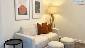 A modern living room with a grey sofa, two brown cushions, a small black table, a floor lamp, and a window with blinds. - The Kode | 2 Bedroom | Family Friendly Home (Drumheller)