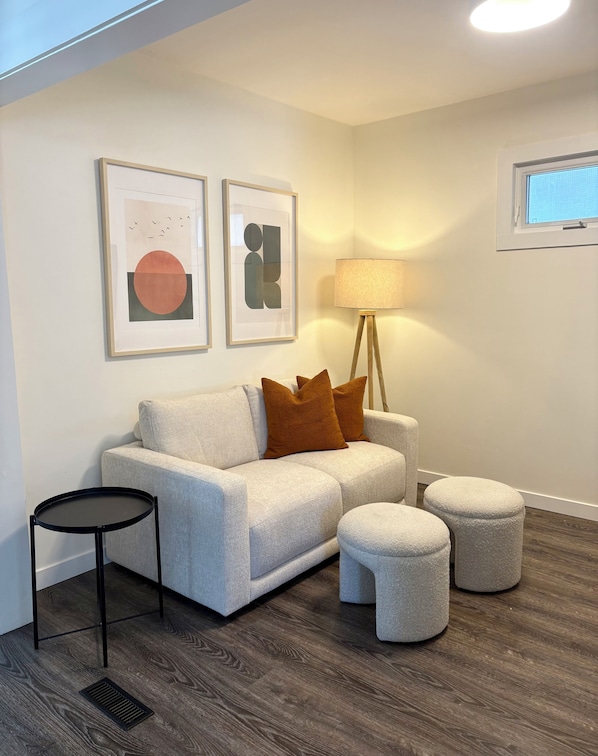 A modern living room with a grey sofa, two brown cushions, a small black table, a floor lamp, and a window with blinds.