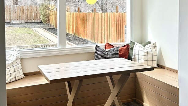 A room with a built-in bench and table by a large window, offering a view of a backyard with a wooden fence and trees. - The Kode | 2 Bedroom | Family Friendly Home (Drumheller)