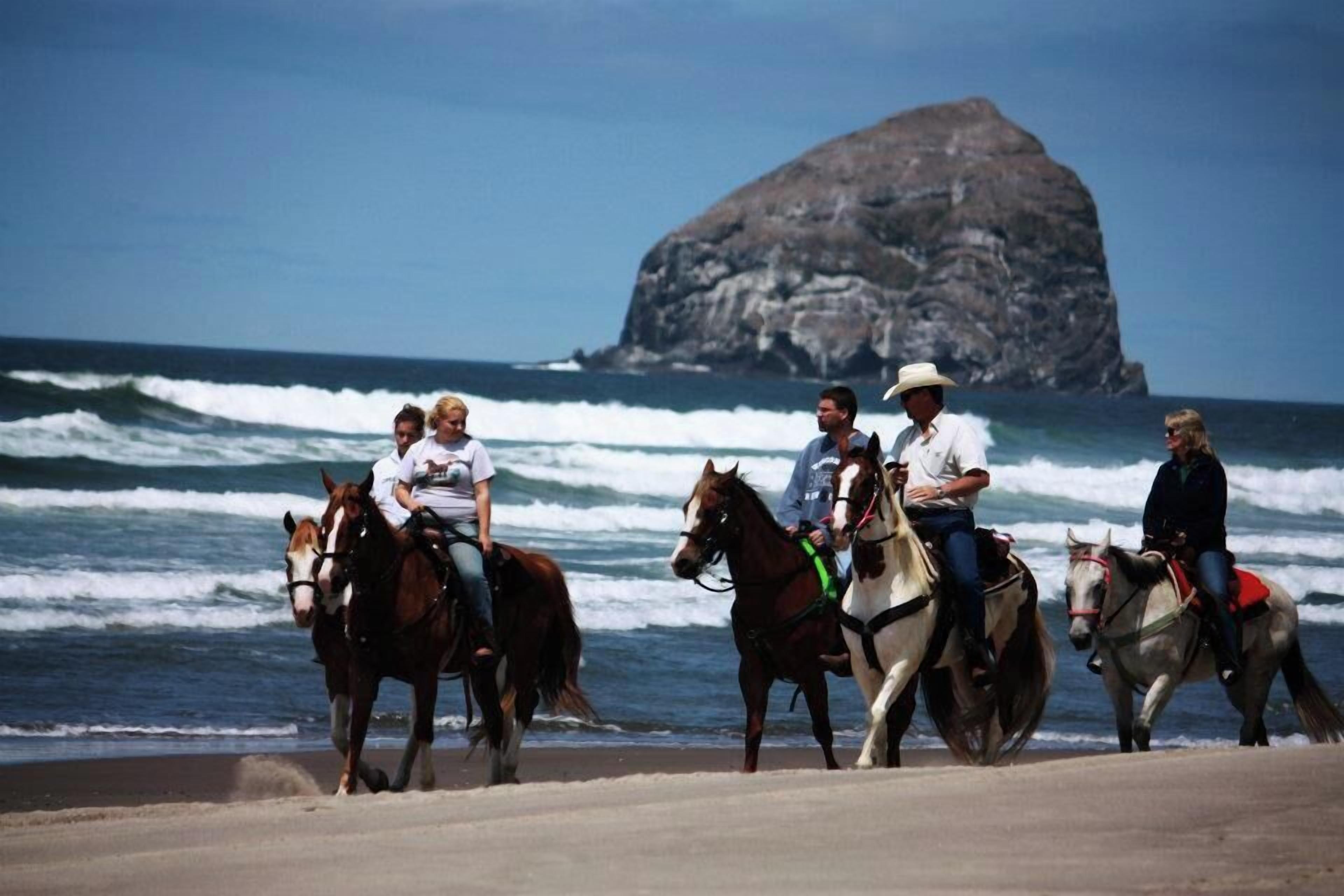 Eine Gruppe von Menschen reitet Pferde am Strand entlang, im Hintergrund sind Meereswellen und eine große Felsformation zu sehen.