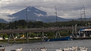 A bridge over water with ducks and a boat, a mountain in the background, and a gas station on the right. - Hostal Irma (Pucon)