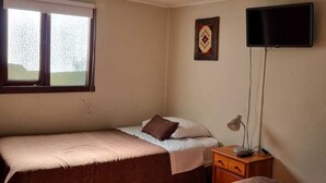 A room with two beds, a TV, a desk with a lamp, and a window with blinds. - Hostal Irma (Pucon)