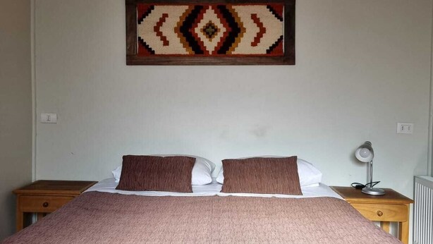 A bedroom with a bed, two nightstands, a lamp, and a wall hanging. - Hostal Irma (Pucon)