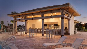 A covered outdoor seating area with chairs and tables, a pool, and a building with a tiled roof. - ChampionsGate Luxury 8BR Villa, Pool, Theater, Game Room (Four Corners)