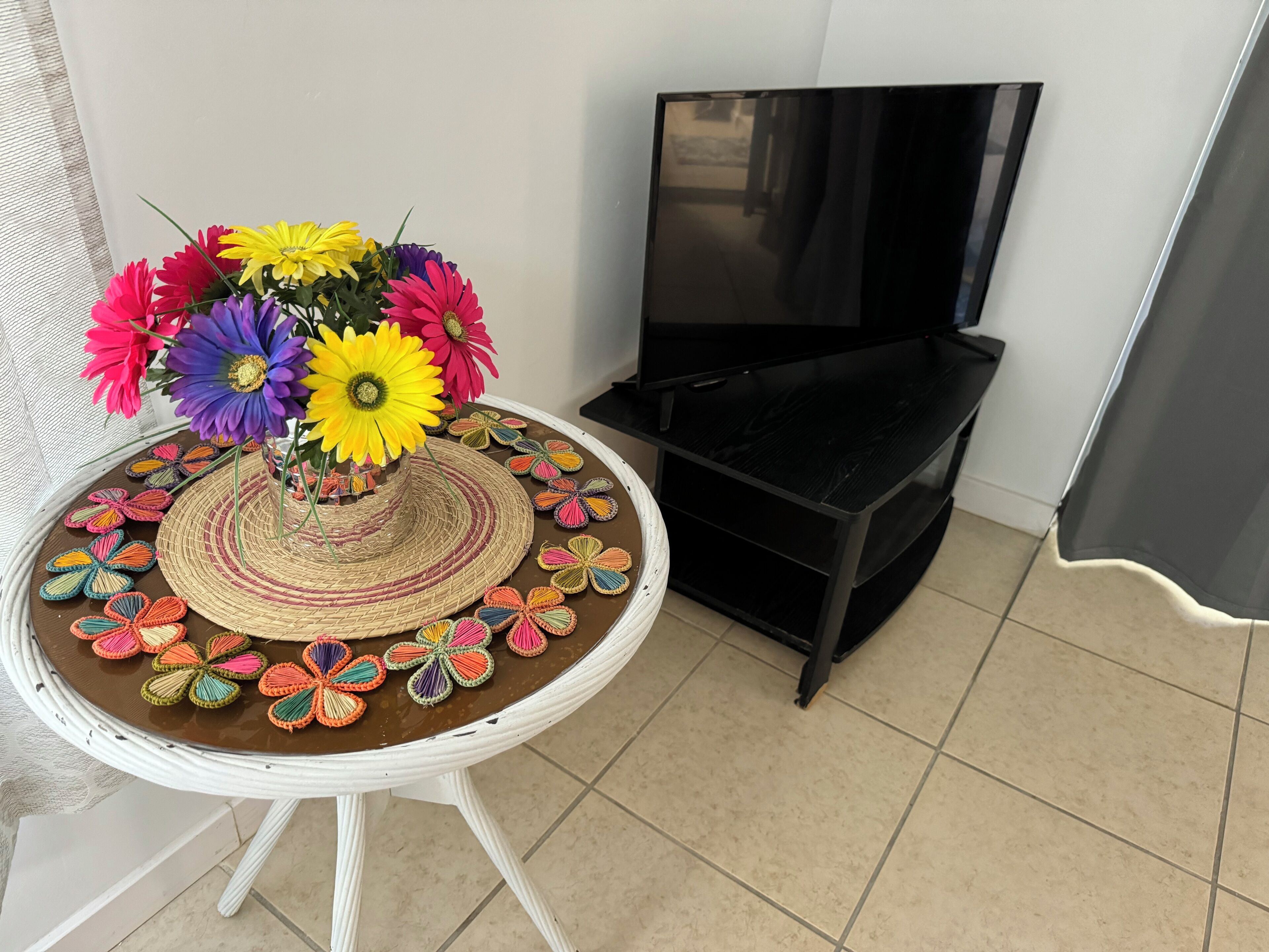 Una mesa redonda con un centro de mesa tejido y flores de ganchillo coloridas, un mueble bajo para televisor negro y un suelo de baldosas.
