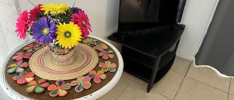 A round table with a woven centerpiece and colorful crocheted flowers, a black TV stand, and a tiled floor.