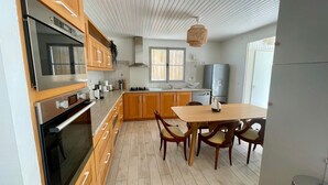 A modern kitchen with wooden cabinets, a built-in oven, a dining table with chairs, and a large window providing natural light. - La Maison Mi Pena - Arcachon basin (Arcachon)