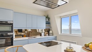 A modern kitchen with light blue cabinets, a central island, and a view of the sea through a window. - No 10 Croft House (Tenby)