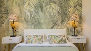 A bed with a green bedspread, two white nightstands, two lamps with polka dot shades, and two pillows with a tropical print.