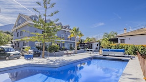 A swimming pool with a tiled deck, surrounded by a white building with a tiled roof, a parked car, and a palm tree.