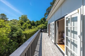 Une terrasse en bois offrant une vue sur une végétation luxuriante, un bâtiment blanc doté de grandes fenêtres et une porte ouverte sur une pièce où se trouvent un lit et une chaise.