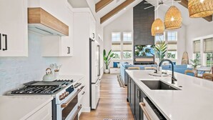 A modern kitchen with white cabinetry, a central island with a sink, and a large window providing a view of the outdoors. - Dune Alright 206 PLA (Port Aransas)