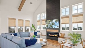 A modern living room with a sectional sofa, a fireplace, large windows with blinds, and a view of palm trees. - Dune Alright 206 PLA (Port Aransas)