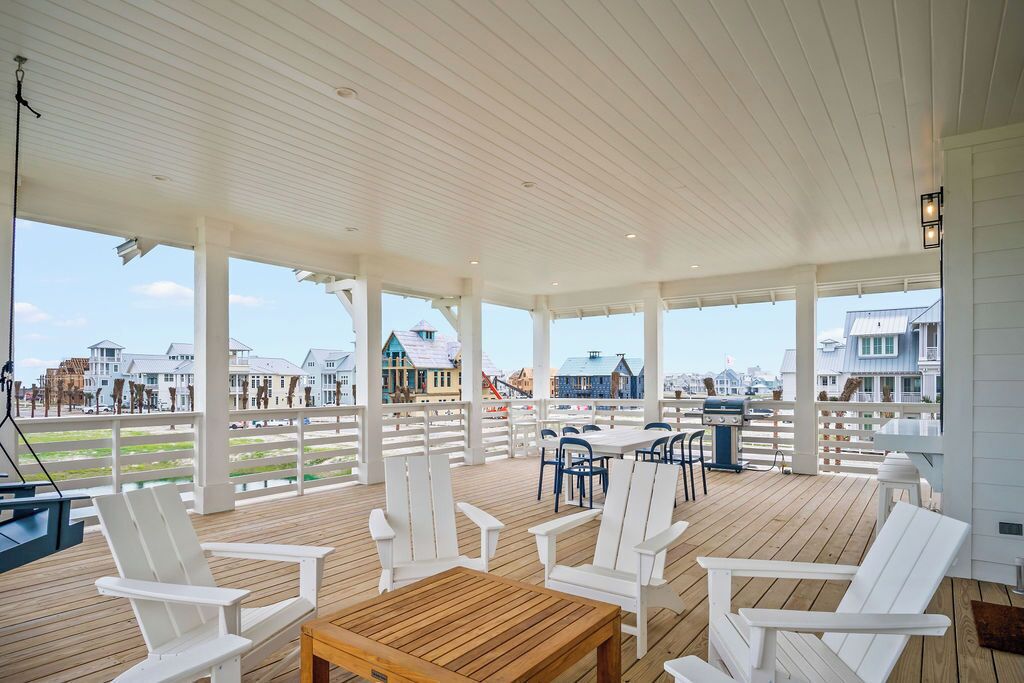 A covered outdoor deck with white chairs and tables, overlooking a waterfront with buildings.