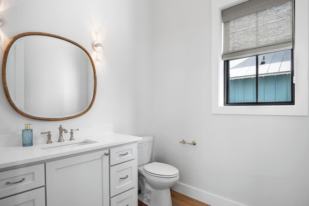 Une salle de bain avec un meuble-lavabo blanc, un miroir rond, des toilettes et une fenêtre avec des stores.