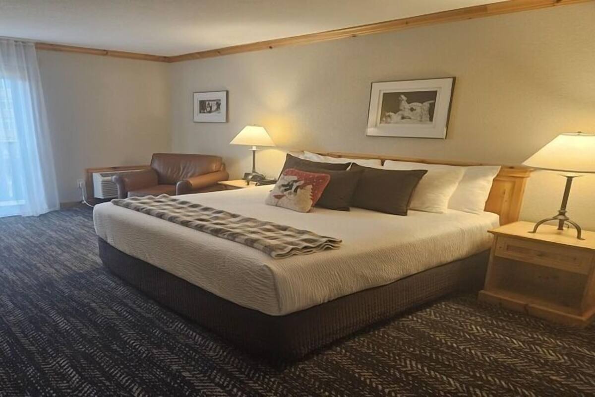 A hotel room with a large bed, two bedside tables with lamps, a leather armchair, and a carpeted floor.