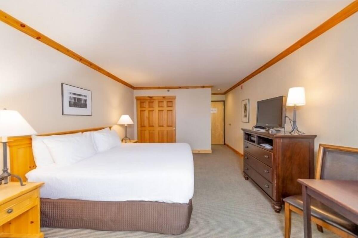 A hotel room with a bed, bedside tables, a dresser, a television, and a door leading to another room.