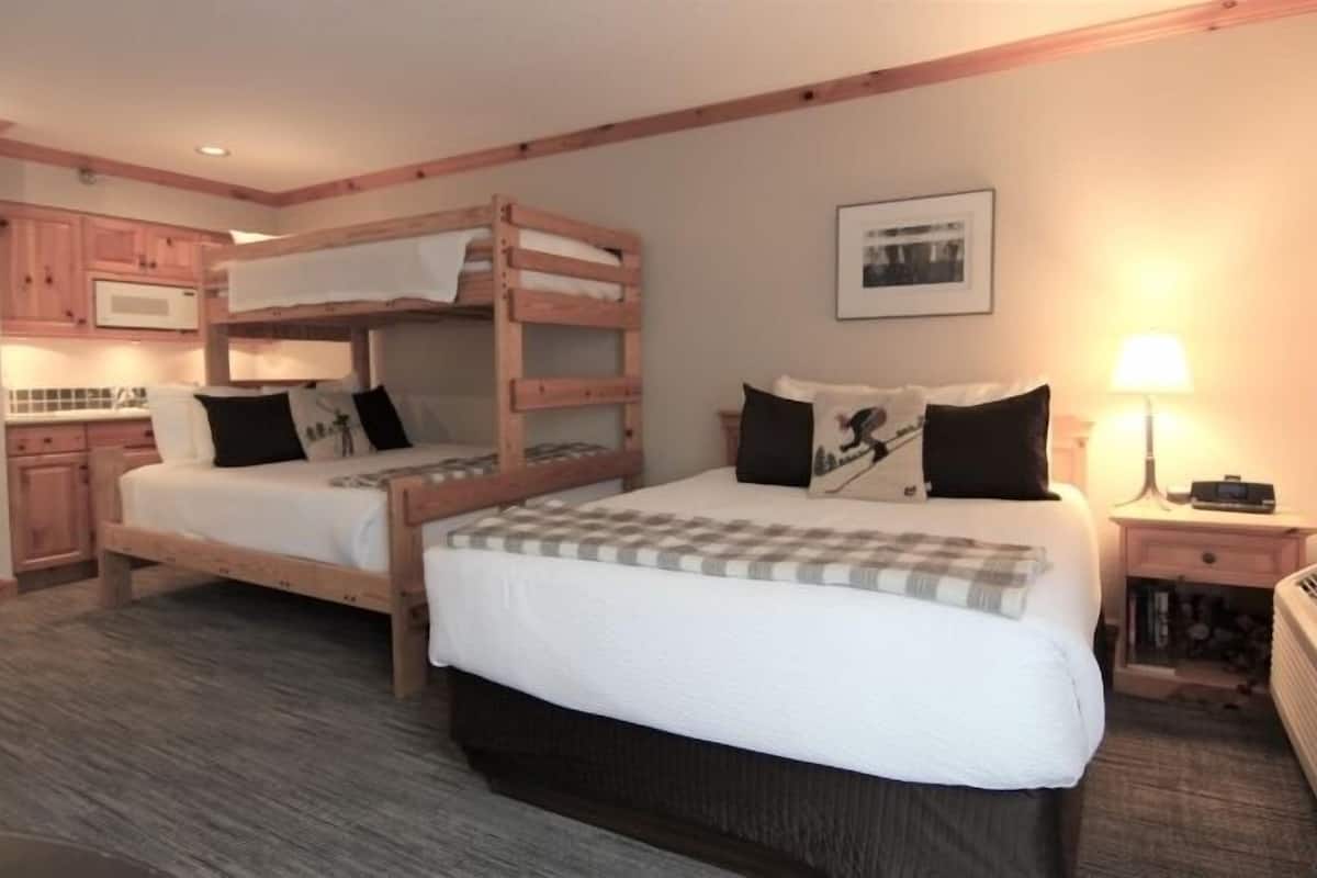 A room with two bunk beds, a small kitchenette, a bedside table with a lamp, and a framed picture on the wall.
