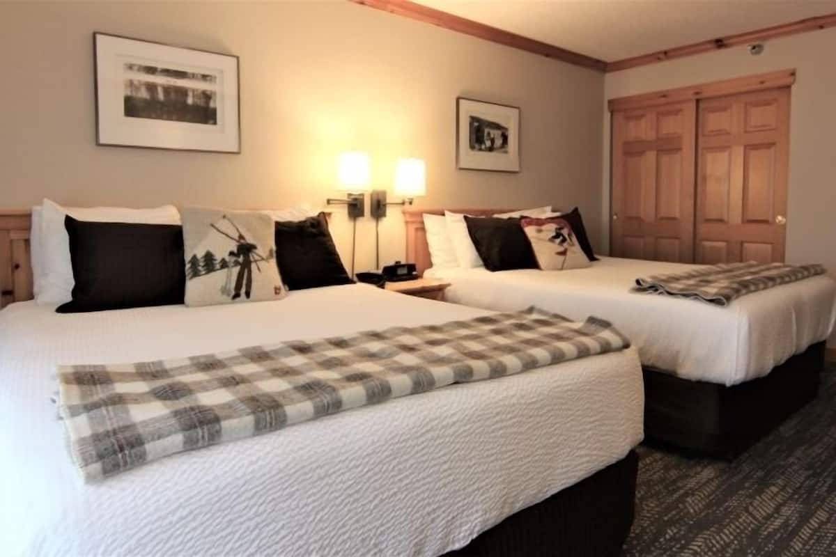 A hotel room with two beds, each with a white comforter and a plaid throw blanket. There are two wall-mounted lamps and two framed pictures above the beds. The room has a wooden door and a carpeted floor.