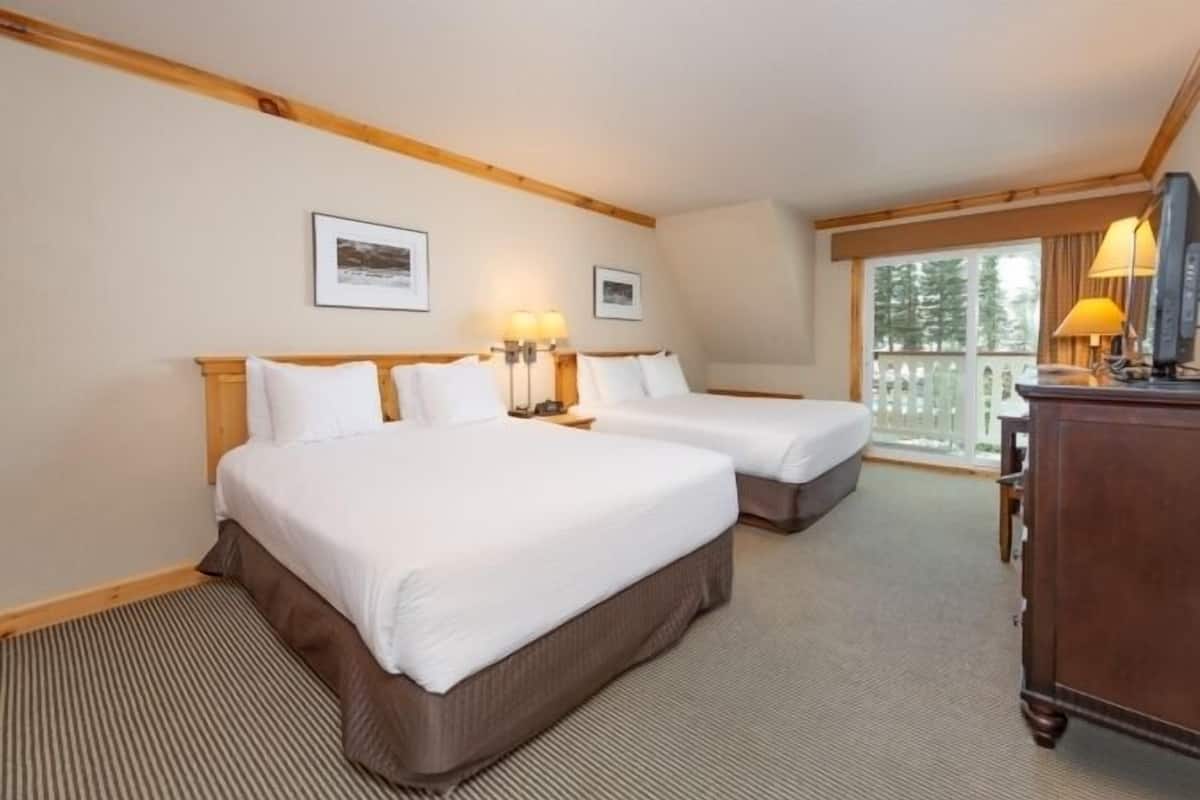 A hotel room with two beds, a wooden headboard, a desk with a lamp, and a window with a view of trees.