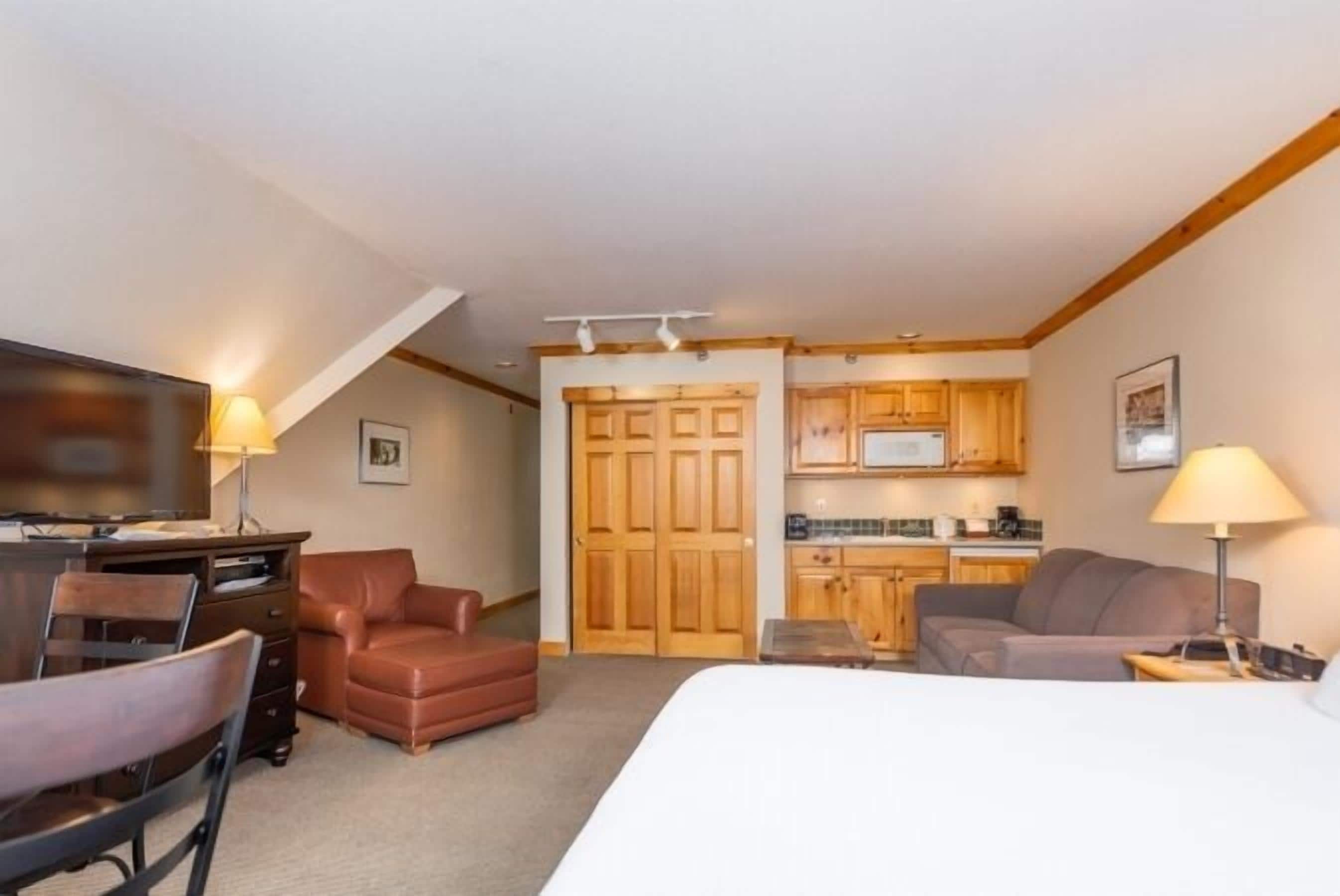Habitación de hotel compacta con cama, escritorio y silla, sofá, pequeña cocina y armarios de madera.