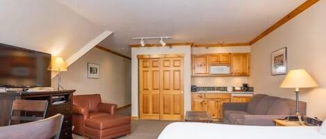 A compact hotel room with a bed, a desk with a chair, a sofa, a small kitchen area, and wooden cabinets.