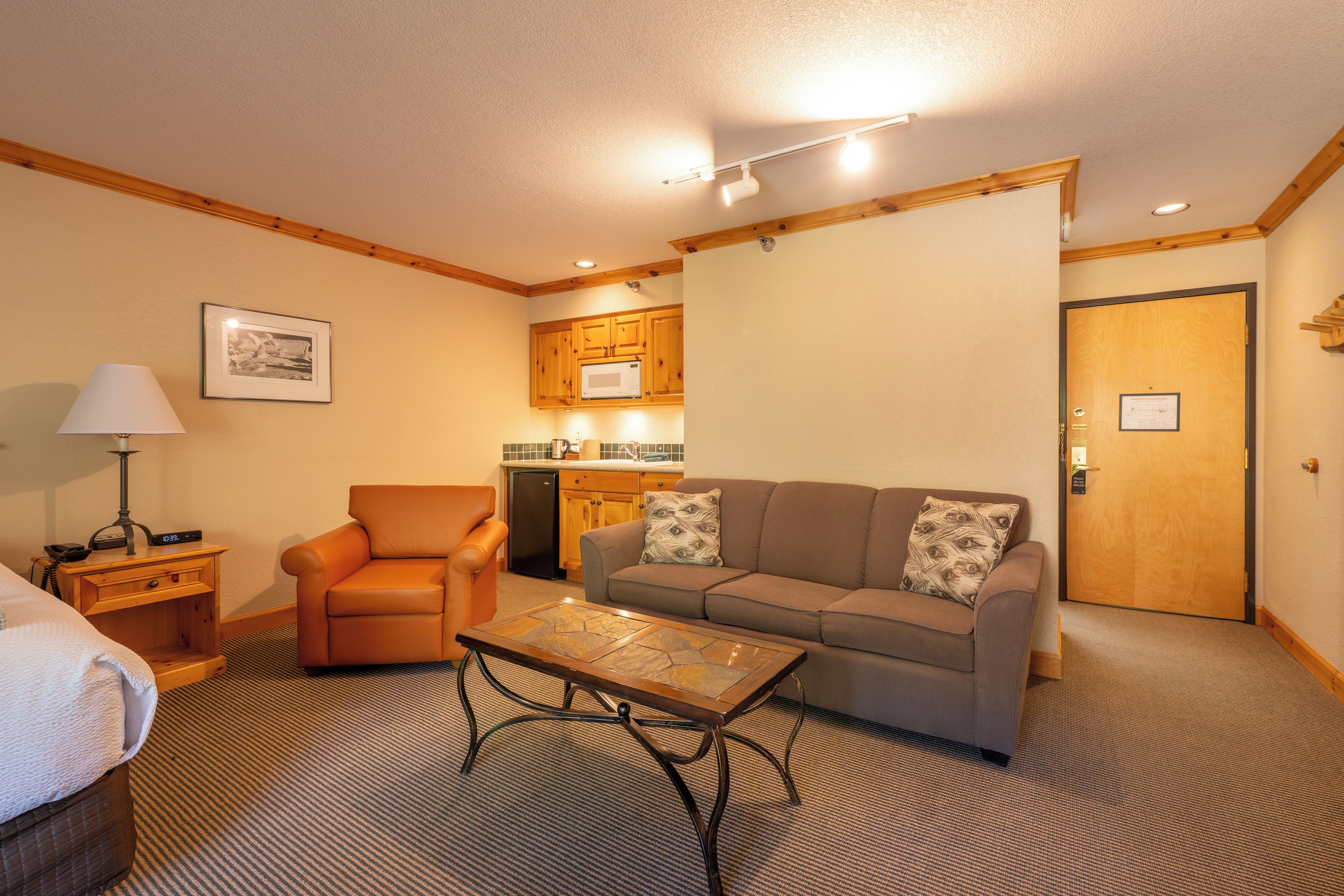 A hotel room with a sofa, armchair, coffee table, kitchenette, and a door.