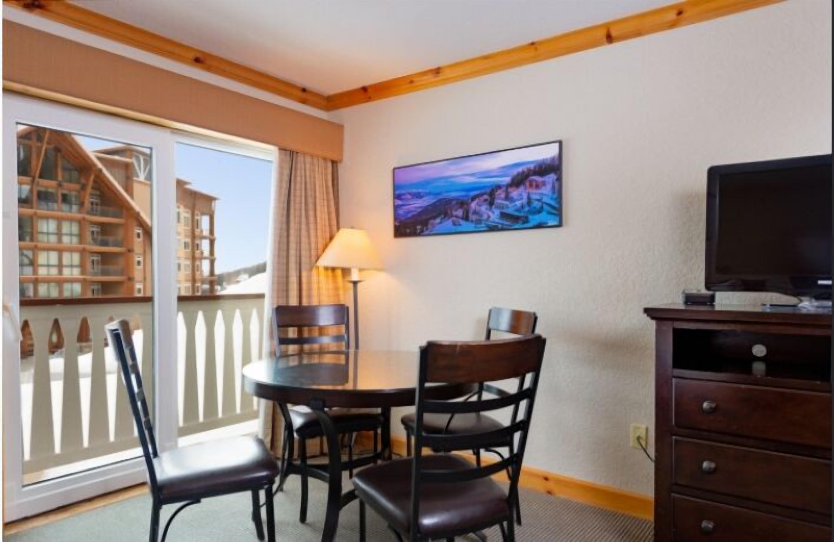 A room with a balcony, a table with chairs, a lamp, a TV, and a painting on the wall.
