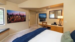 Habitación de hotel con cama, escritorio con computadora, televisor y un cuadro de un paisaje montañoso.
