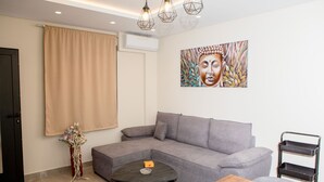 Ein modernes Wohnzimmer mit einer grauen Eckcouch, einem kleinen runden Couchtisch und einem Holztisch. An der Wand hängt ein Gemälde, und auf einem Podest steht eine Vase mit Blumen.