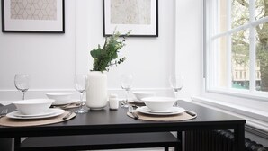 A dining table set for four with white plates, bowls, and wine glasses, a white vase with greenery, and framed artwork on the wall.