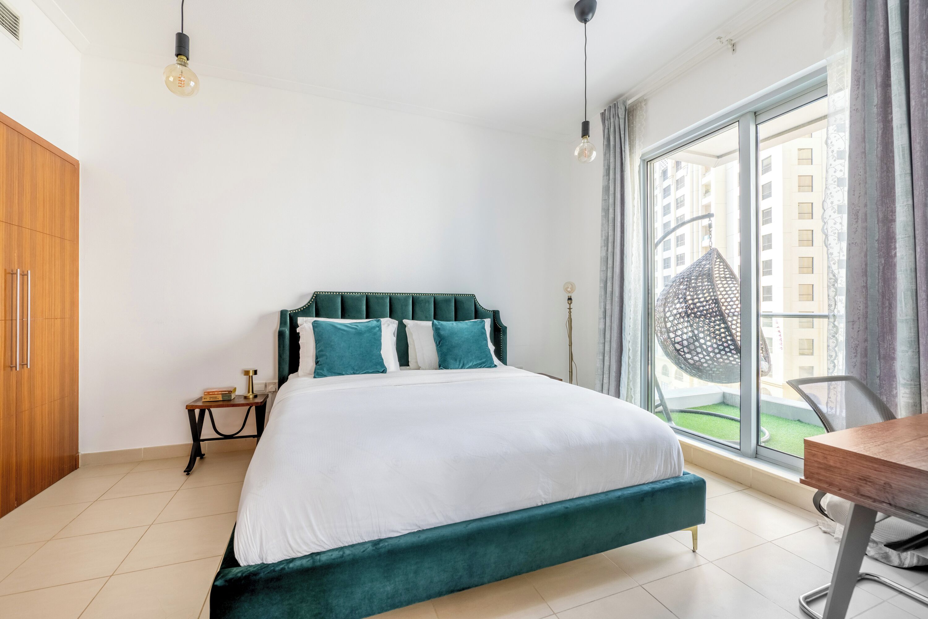 A modern bedroom with a large bed, a wooden wardrobe, a small bedside table, and a view of a cityscape through a large window.