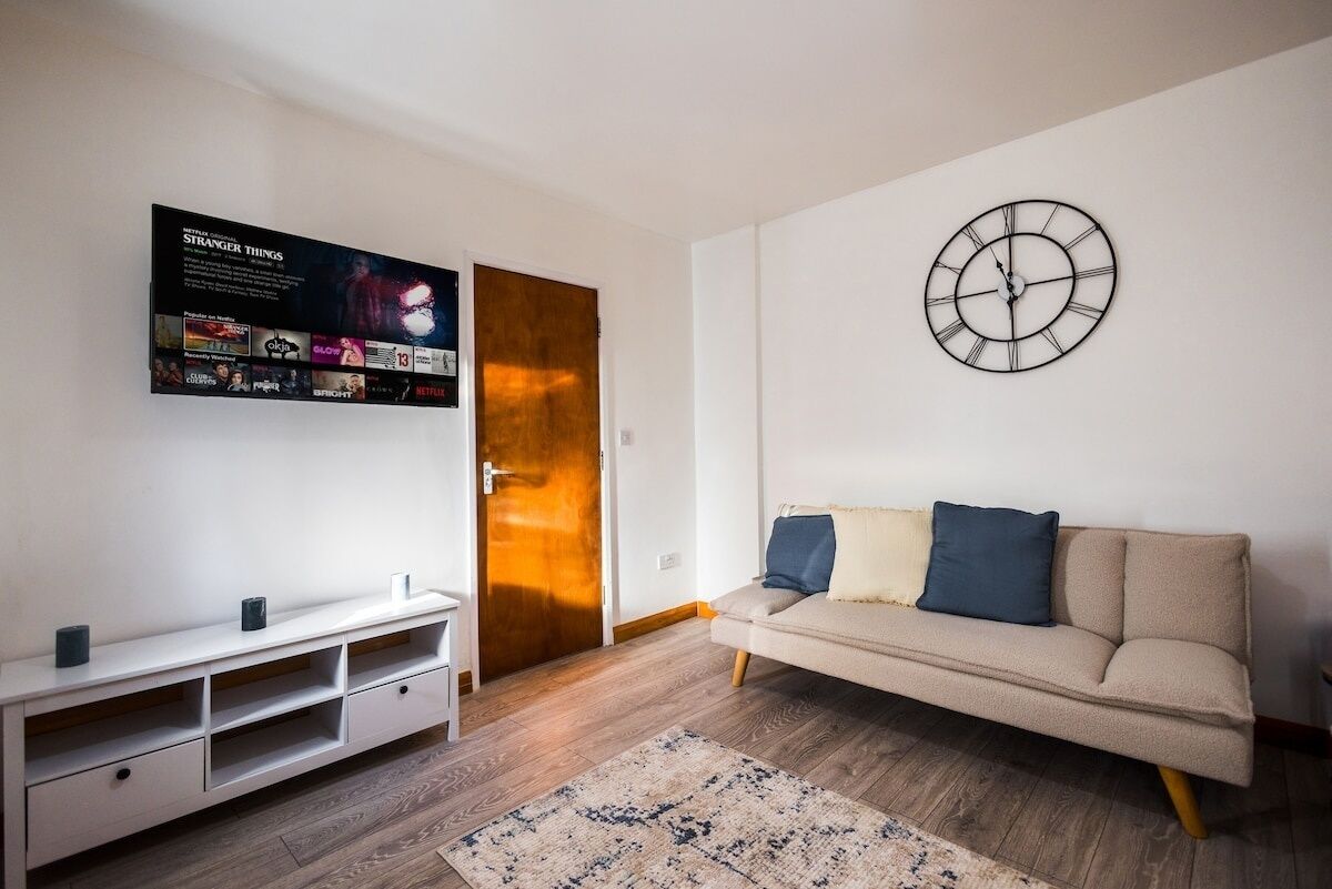 Una sala de estar con un sofá, un televisor montado en la pared, una puerta de madera y un reloj.