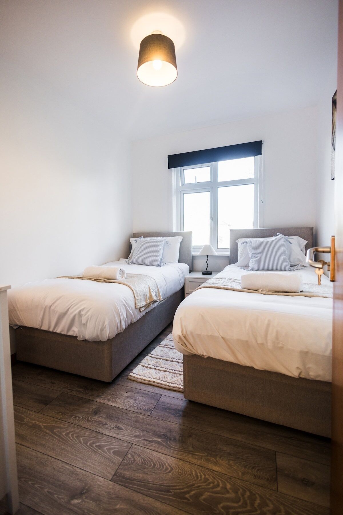 A bedroom with two beds, a window with a view, a lamp, and a wooden floor.