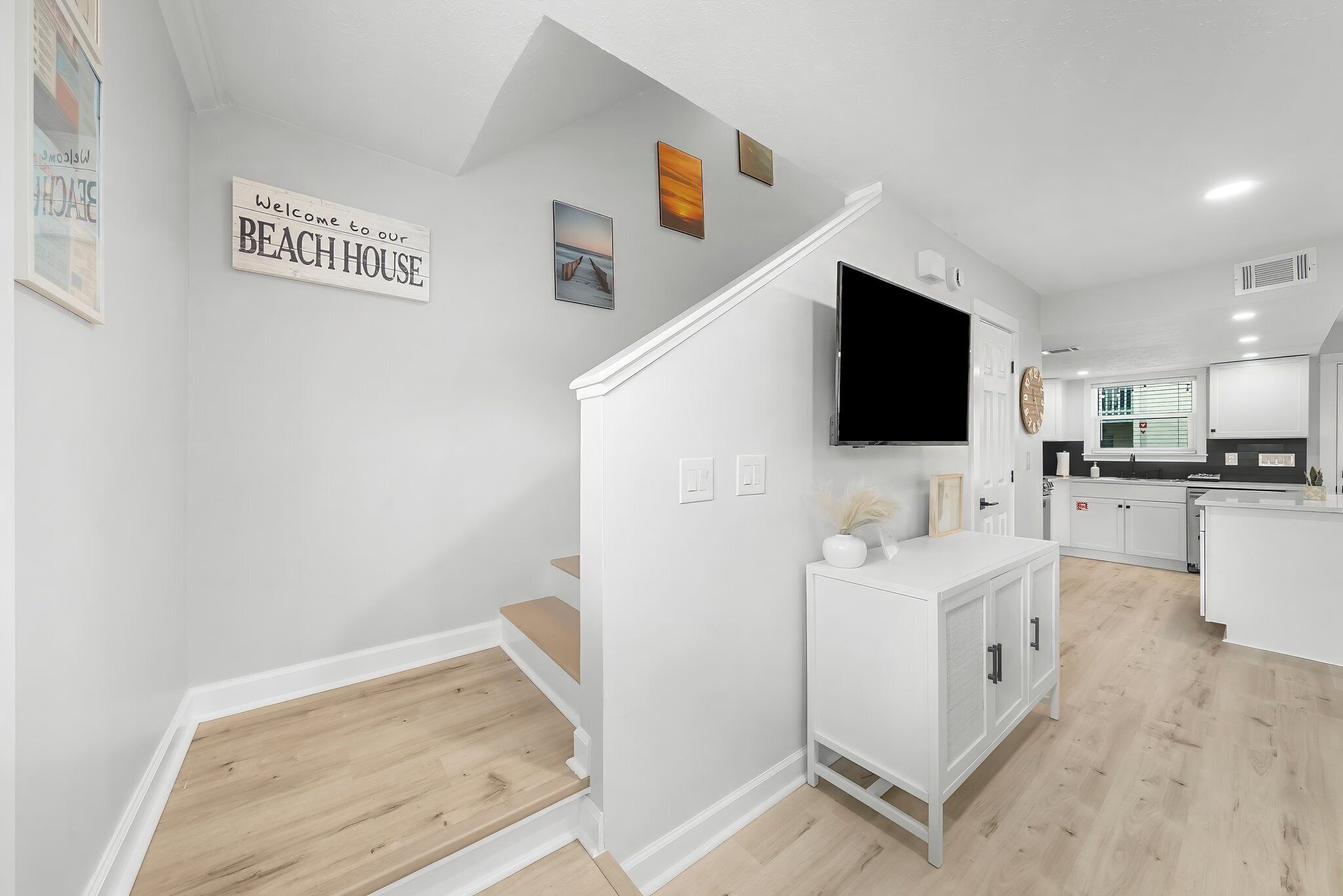 A modern kitchen with white cabinetry, a flat-screen TV mounted above the counter, and a wooden staircase leading to another level.