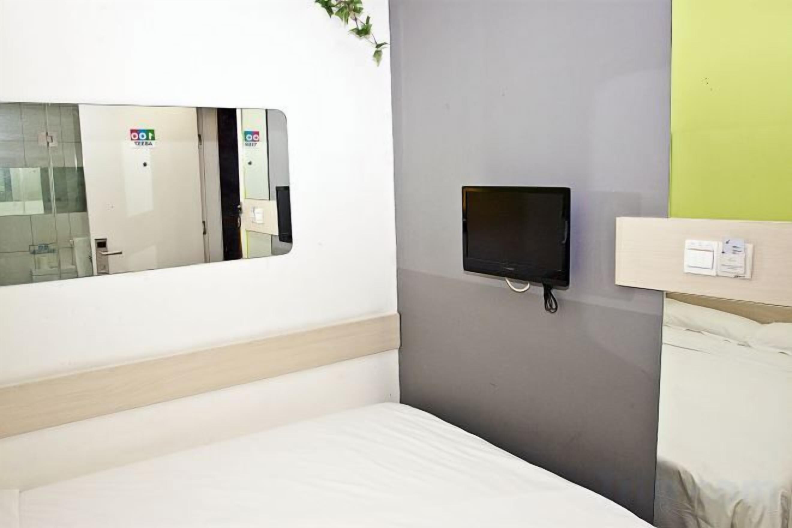 A hotel room with a bed, a small TV mounted on the wall, and a window with a reflection of a door and a sign.