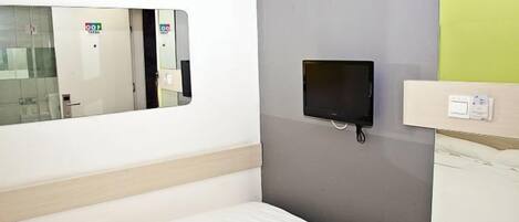 A hotel room with a bed, a small TV mounted on the wall, and a window with a reflection of a door and a sign.