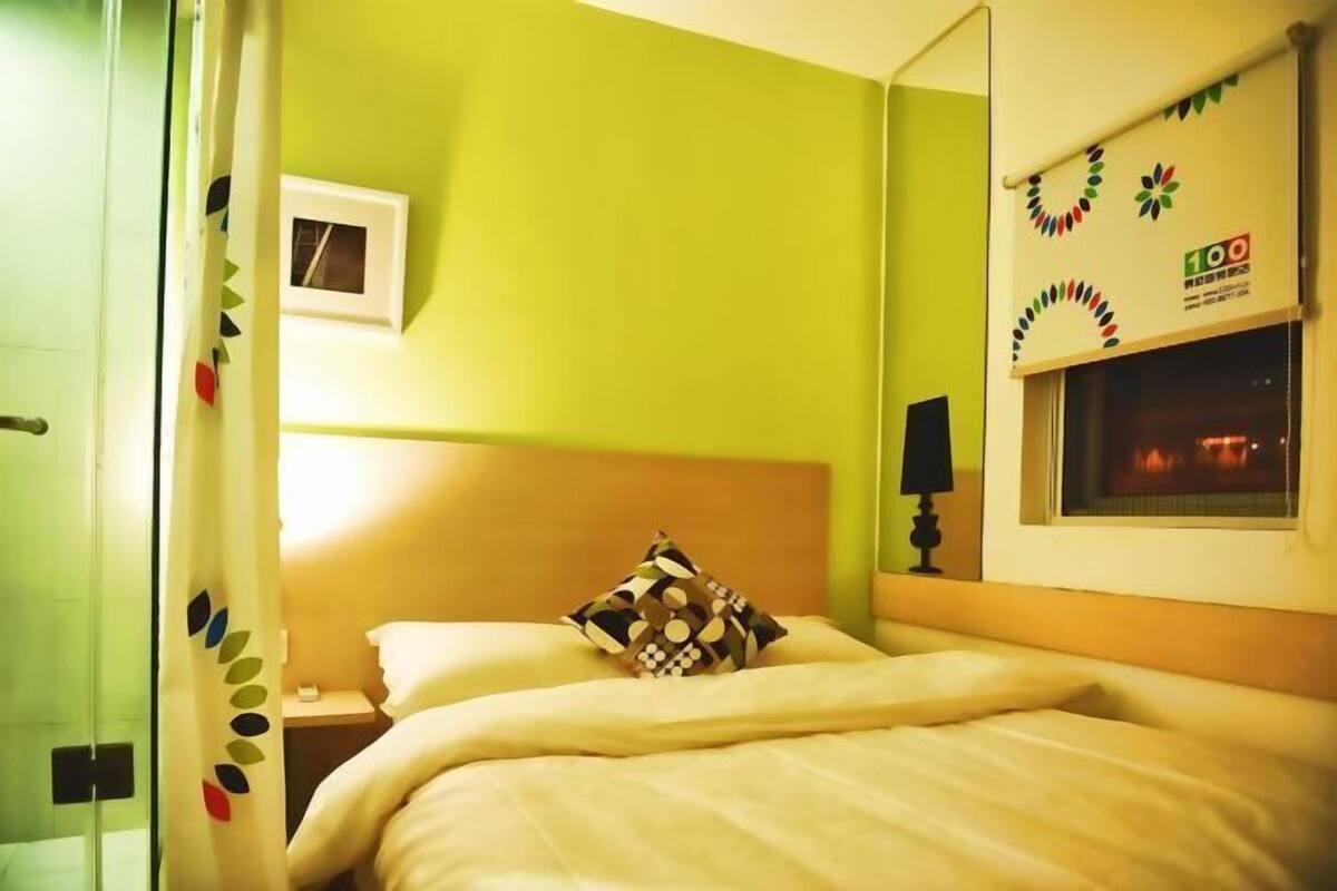Una habitación de hotel con cama, mesita de noche, lámpara, ventana con vistas y espejo en la pared.
