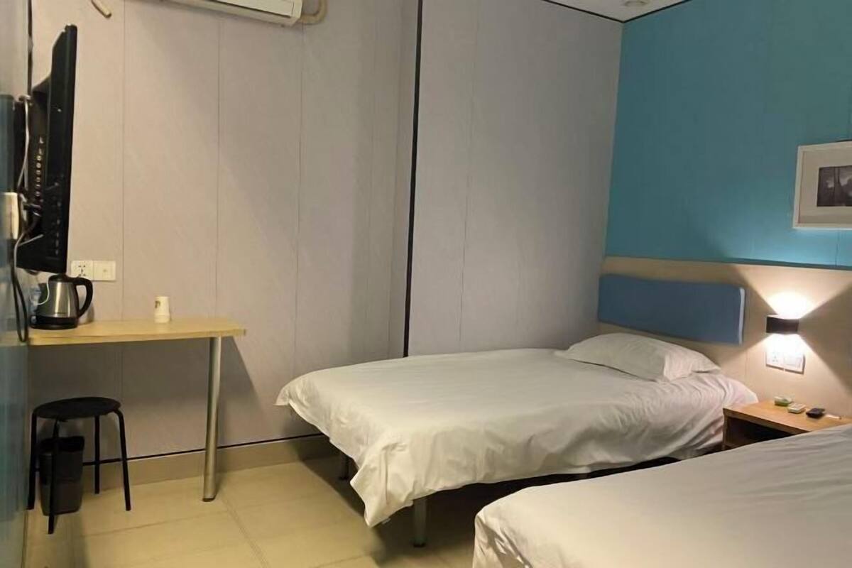 Habitación de hotel con dos camas, un escritorio con hervidor, una silla y una mesita de noche con lámpara.