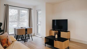 En moderne stue med et fladskærms-tv på et træstel, et spisebord med stole, og en sofa med en hundepude.